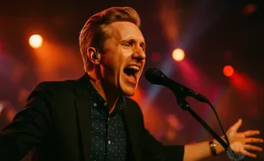 Man singing into a microphone on stage, with red and orange stage lights.