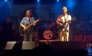 A band performs on stage, with two men in the foreground playing acoustic guitars and singing, and a drummer in the background.