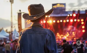 Person in cowboy hat and denim jacket facing a brightly lit outdoor concert stage at sunset.
