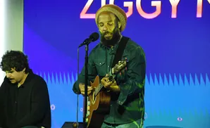 A man with a beard and a tan hat plays an acoustic guitar and sings into a microphone, with another man sitting to his left.