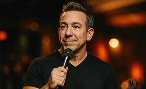 A man in a black shirt holds a microphone and smiles at a camera.