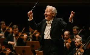 An elderly male conductor with white hair smiles, holding a baton and leading an orchestra.