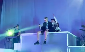 Singer in a suit and shorts sits on a platform holding a microphone, while another person behind him raises their arm.
