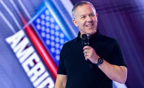 A man in a black shirt holds a microphone and smiles against a backdrop of the American flag.