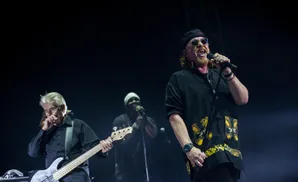 Singer with a red mustache and sunglasses performs onstage with a bassist and backup singer.