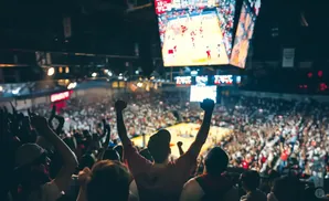 Fans cheer at a basketball game, with a large screen showing the court.