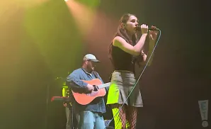 A female singer holds a microphone while a male guitarist plays an acoustic guitar.