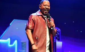 A Black man in a red shirt and pants smiles while holding a microphone on a blue-lit stage.