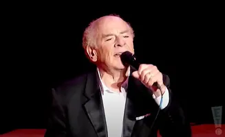 An older man in a suit and white shirt sings into a microphone on a dark stage.