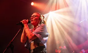 A female singer with glasses and tattoos performs on stage, illuminated by stage lights.