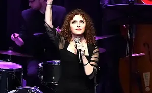 Bernadette Peters sings into a microphone with her arm raised on stage.