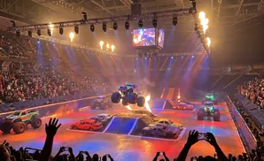 Monster truck show with trucks jumping over cars, flames, and a large crowd watching.