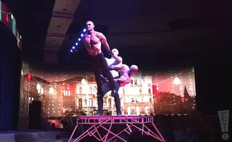 Three muscular men perform a balancing act on a stage, with a painted cityscape backdrop.