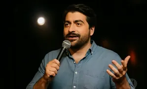 A man in a blue shirt holds a microphone and gestures with his left hand while speaking.