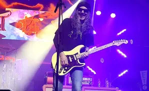 A guitarist with long hair and sunglasses plays a cream-colored Fender Stratocaster on a stage lit by purple lights.