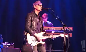 A guitarist sings into a microphone while another musician plays keyboards behind him.