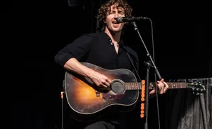 Musician with curly hair sings into a microphone while playing an acoustic guitar.