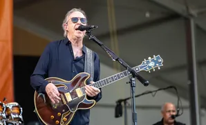 A man in sunglasses plays a brown hollow-body guitar on stage while singing into a microphone.