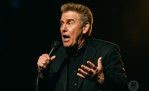 Comedian Brian Regan holds a microphone and gestures with his hands while speaking on stage.
