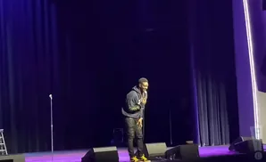 A man in a hoodie and leather pants laughs on a stage with purple curtains.