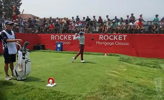 Golfer at Rocket Mortgage Classic swings a club while a caddie stands by a golf bag, with spectators in the background.