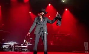Singer in a black fedora and embellished suit performing on a red-lit stage, holding a dark cloth.