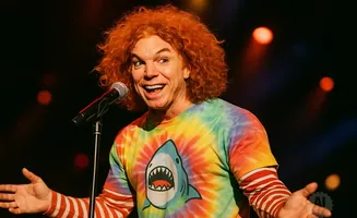 Comedian Carrot Top with bright orange curly hair, wearing a tie-dye shirt with a shark graphic, stands at a microphone on stage.