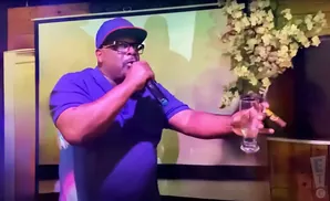 A man in a purple shirt and blue hat holds a microphone and a cigar, with a glass of liquor.