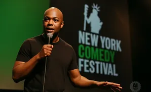 A bald Black man in a black t-shirt holds a microphone and gestures with his other hand in front of a 