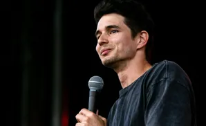A man smiles while holding a microphone, wearing a dark t-shirt against a dark background.