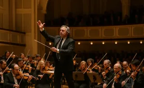 An orchestra conductor with his arm raised, leading musicians playing violins in a concert hall.