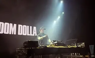 A DJ wearing a black shirt and hat plays music on stage with the name 