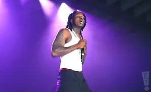 A man with dreadlocks and tattoos, wearing a white tank top and black pants, sings into a microphone on a purple-lit stage.