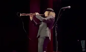 A musician in a hat and suit plays a saxophone on a dimly lit stage.