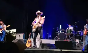 A country band performs on a stage with a singer in a white cowboy hat and mask playing a white guitar.
