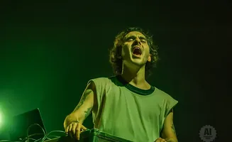 A man with curly hair sings with his mouth open, wearing a sleeveless shirt and standing behind DJ equipment.