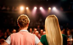 Backs of a man and woman facing a dimly lit audience, spotlights overhead.