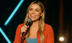 A blonde woman in an orange jacket smiles while holding a microphone.