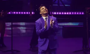 A man in a purple suit and polka-dot tie claps enthusiastically on a dimly lit stage with purple lights.