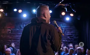 Man with microphone on stage, facing away from camera, with audience in the dark background.