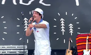 A man in white overalls and a bucket hat sings into a microphone on a stage with a dark backdrop and white tree graphics.