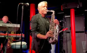 Man plays a saxophone on stage with a keyboardist in the background.