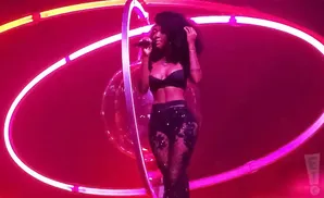 Singer performs on stage wearing a black bra and sheer sparkly pants, illuminated by pink and yellow neon lights.