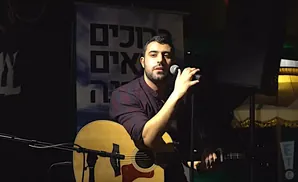 A man with a beard sings into a microphone, playing an acoustic guitar on stage.