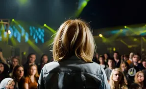 Woman with blonde hair facing away from camera on stage at a concert with yellow stage lights and a blurred crowd.
