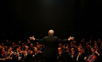 A conductor directs an orchestra from behind, with arms outstretched against a dark background.
