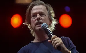 A man with blonde hair and a mustache speaks into a microphone, with red and blue lights behind him.