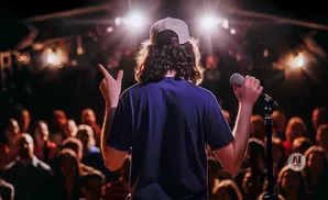 A man with long curly hair and a backward baseball cap stands on a stage, holding a microphone and gesturing to a blurry audience.