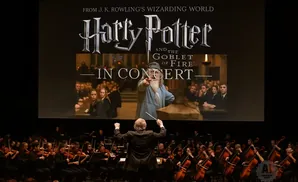 A conductor leads an orchestra playing in front of a screen showing 