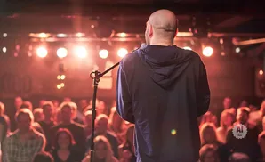 A bald man in a hoodie on stage with a microphone, facing a blurry audience under warm stage lights.